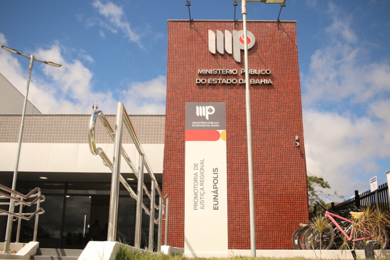 Prédio branco com coluna de tijolos vermelhos e placa escrito "Promotoria de Justiça Regional de Eunápolis"